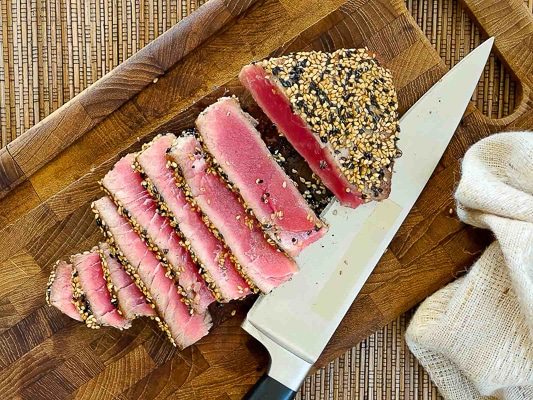 Seared ahi tuna being sliced on a wooden cutting board with a chef's knife on the side.