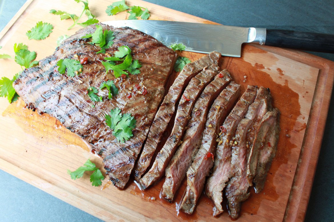 sliced flank steak topped with fresh cilantro on a wooden cutting board with a chef's knife on the side.