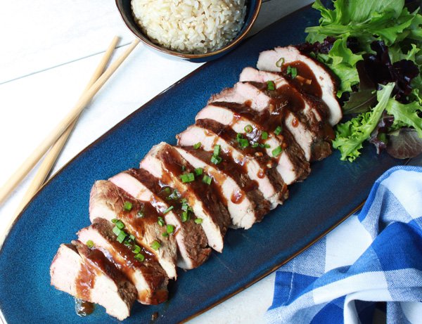 sliced pork tenderloin on a blue place with a side of brown rice and chopsticks
