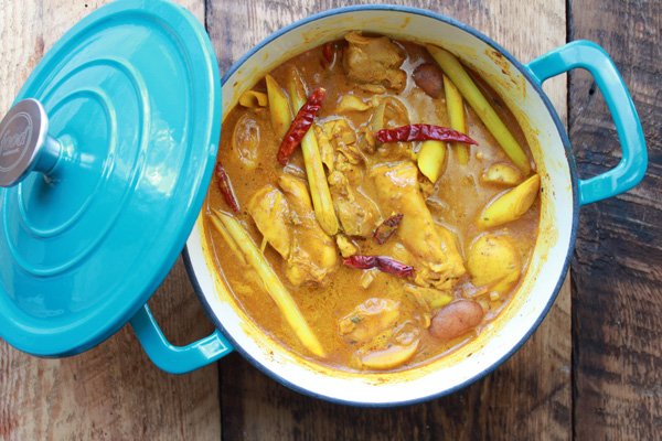 A blue stock pot filled with Chinese chicken curry and potatoes topped with spicy red peppers and stalks of lemon grass on top of a wooden board.