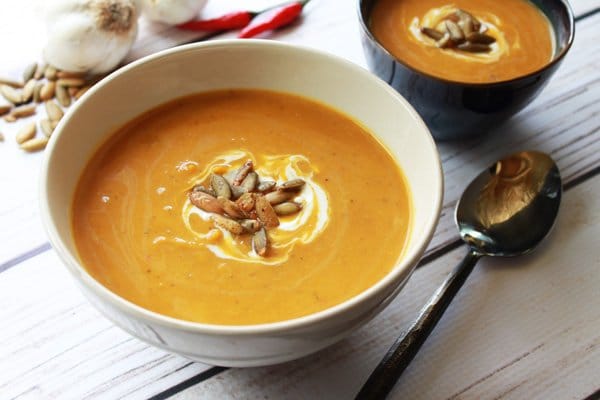 A white soup bowl filled with pumpkin coconut soup topped with toasted pumpkin seeds and swirls of sour cream with a soup spoon on the side and a smaller soup bowl in the background.