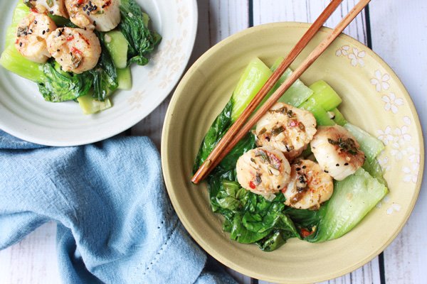 Two plates of seared scallops over a bed of baby bok choy and chopsticks with a blue napkin on the side.