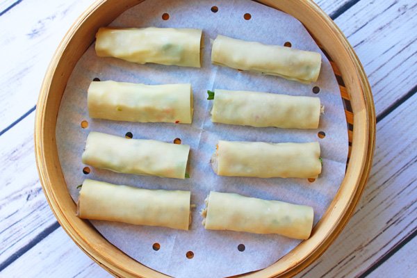 Uncooked steamed Vietnamese rolls on top of parchment paper inserted into a bamboo steamer on top of a white wooden board.