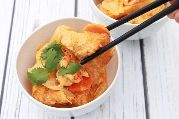 A white bowl filled with coconut curry tofu and shrimp with black chopsticks picking up a piece of tofu on top of a white wooden board.