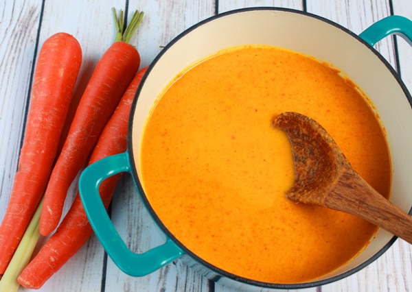 A blue stock pot of cold carrot soup on a white plank wooden board with fresh carrots on the side.