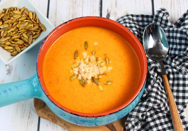 a blue soup bowl with handle filled with cold carrot soup topped with toasted pumpkin seeds and coconut flakes with a soup spoon on the side on top of a black and white checkered napkin.