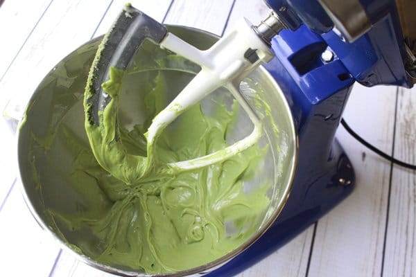 A blue stand mixer with matcha cheesecake muffin batter in the bowl and batter on the beaters on a white plank wooden board.