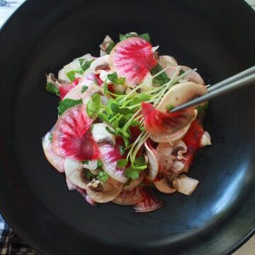 A vibrant mushroom and radish salad in a black bowl with sprouts on top and silver chopsticks on the side.