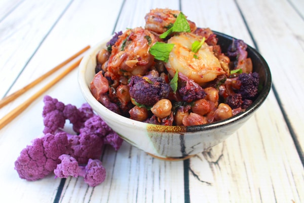 A black and white bowl filled with shrimp sambal and roasted cauliflower and chickpeas topped with a fresh mint leaf with ginger bulbs on the side on top of a wooden white board.