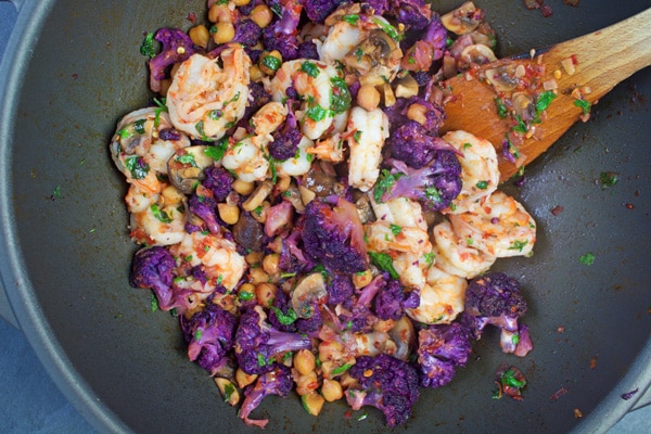 Overhead view of a wok with shrimp, purple cauliflower, chick peas, mint and cilantro being stir-fried with a wooded spatula.