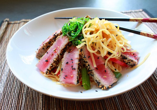 Sesame crusted yellowfin tuna in a white bowl on a bed of Asian noodles with seaweed salad on top and chopsticks on the side on a bamboo placemat.