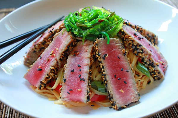 sliced sesame crusted tuna on a bed of Asian noodles topped with seaweed salad in a white bowl with chopsticks on the side