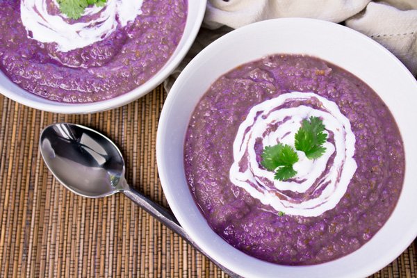 Purple potato and cauliflower soup in two white bowls topped with swirls of sour cream and garnished with sprigs of cilantro with a spoon and beige napkin on the side on top of a bamboo placemat.