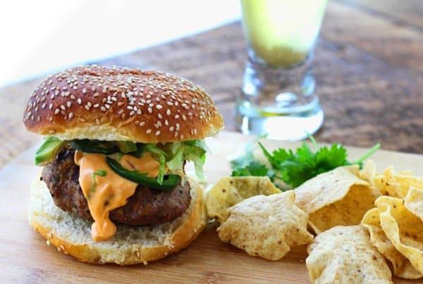 A Thai Turkey Burger topped with Spicy Aioli, lettuce and jalapeno peppers, with tortilla chips on the side with sprigs of bright green cilantro on a wooden board with a glass of beer behind it.