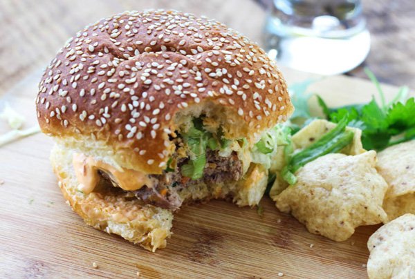 Thai turkey burger with spicy aioli with a bite out of it on to of a wooden cutting board with tortilla chips and sprigs of cilantro on the side.