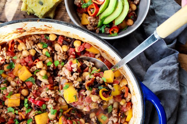 A large blue stock pot of pork chili with colorful beans with a ladle inserted.