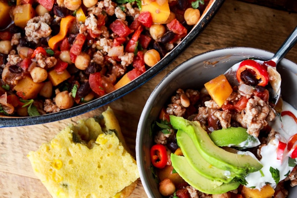 Asian spiced pork chili with butternut squash in a white bowl topped with sour cream and avocado slices with a spoon in the bowl and a square of jalapeno corn bread on the side, and a large pot of chili on a wooden board placed behind it.