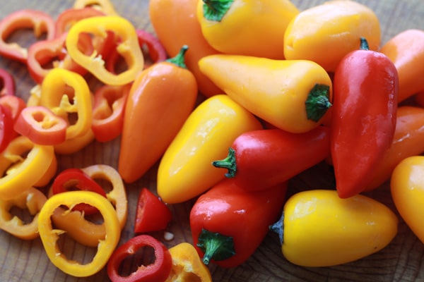 Orange, yellow and red mini bell peppers with sliced peppers on the side on top of a wooden cutting board.