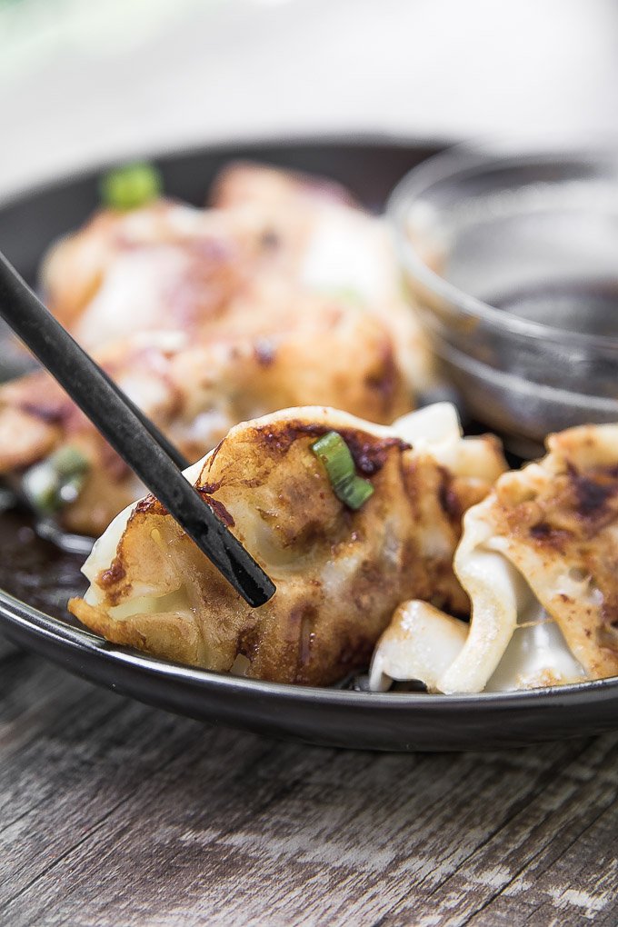 Pork Potstickers held by chopsticks above a black plate with a clear glass bowl of dipping sauce on the side on top of a dark wooden board.a