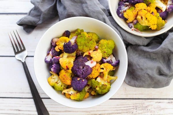 Tri-Colored cauliflower in a white bowl with an orange piece of cauliflower held on a fork with a grey napkin on the side on a wooden board