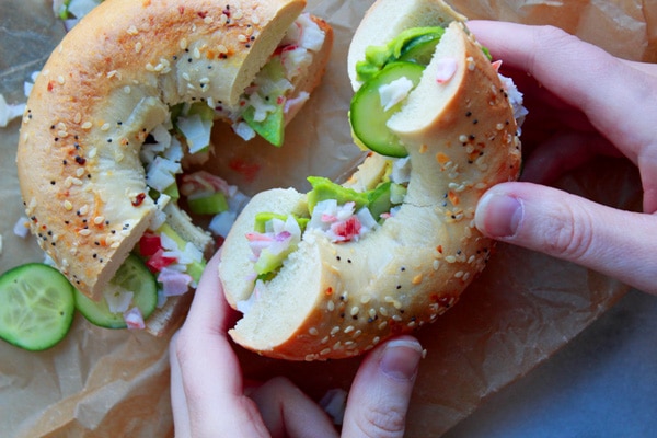 Two hands holding a California Everything Bagel Sandwich cut in half on top of brown parchment paper with a few slices of cucumber on the side.