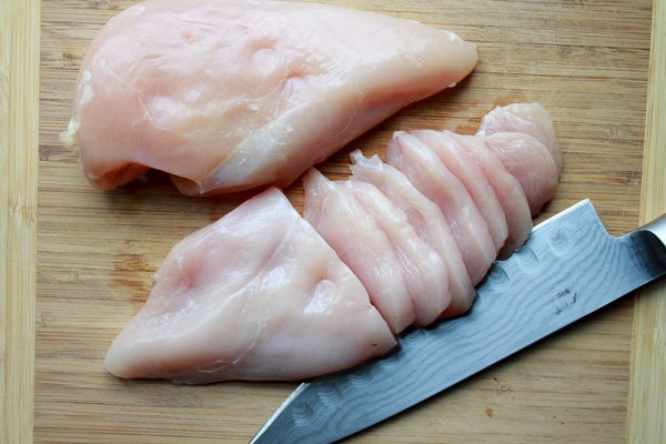 sliced uncooked chicken breasts on a wooden cutting board with a chef's knife on the side
