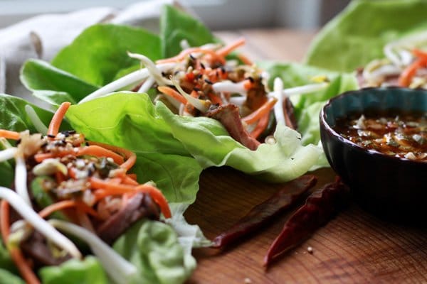 Grilled flank steak lettuce cups on a wooden board with a side of nuoc cham sauce in a dark blue bowl with hot peppers  