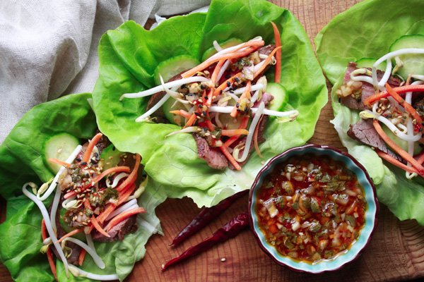 Overhead view of grilled flank steak lettuce cups on a wooden board with a side of nuoc cham sauce in a blue bowl with hot peppers.