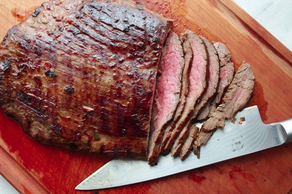 Sliced grilled flank steak with a chef's knife on the side on top of a wooden cutting board.