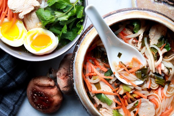 A bowl of miso chicken noodle soup with a white spoon inserted and a smaller bowl of chicken noodle soup behind topped with runny eggs.