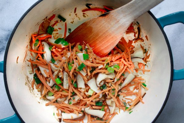 Mixed vegetables sautéing in large blue soup pot with a wooden spatula inserted inside.  