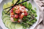 Mixed seafood poke bowl in a white bowl with cucumbers, avocado and radish sprouts surrounding it with lime wedges and chopsticks on the side with a beige napkin on the side