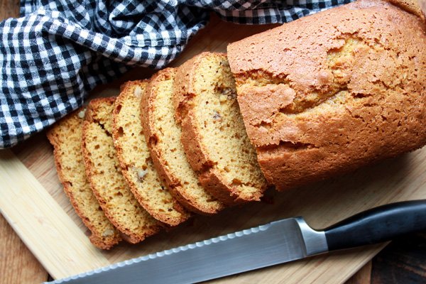 A sliced loaf of five spice pumpkin bread on a wooden board with a bread knife and checkered napkin on the side.