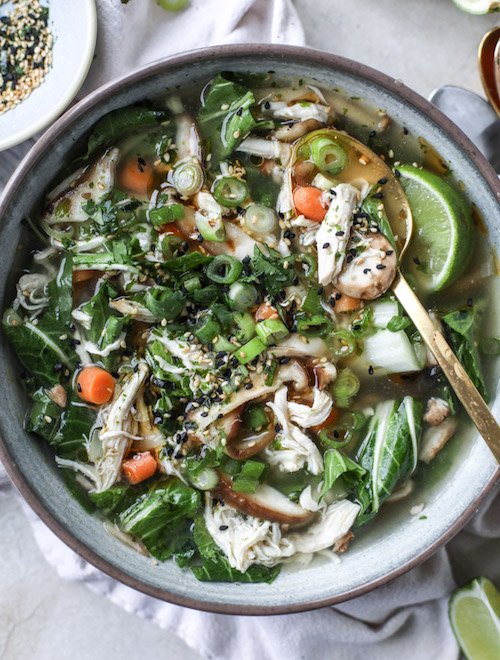 A grey bowl filled with Asian Chicken Soup with lots of fresh green veggies and a spoon inserted on top of a gray napkin.