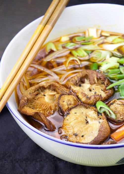 A white bowl filled with Asian bone broth soup topped with shiitake mushrooms and a pair of chopsticks.