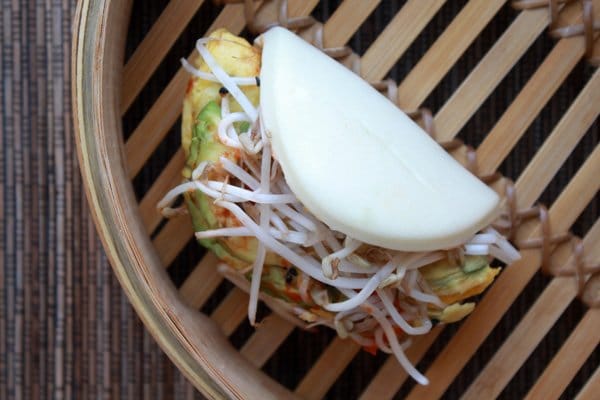 A steamed bao bun stuffed with eggs, avocado slices and bean sprouts sitting inside a bamboo steamer with the lid off.