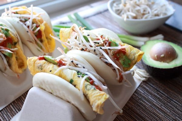 steamed buns with eggs, avocado slices, sprouts, and a sriracha drizzle in a taco holder with avocado and sprouts on the side