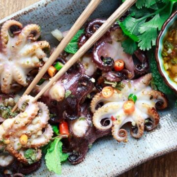 Grilled baby octopus on a long gray serving platter with a small bowl of dipping sauce on the side and a pair of chopsticks.
