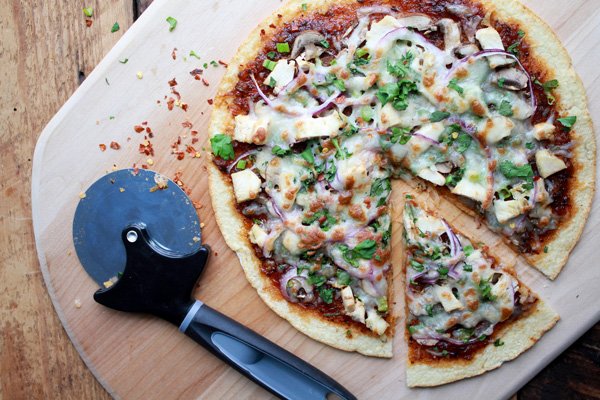 Korean BBQ Cauliflower Pizza pie topped with veggies and fresh herbs on top of a wooden pizza peel with one slice cut out and a pizza cutter on the side.