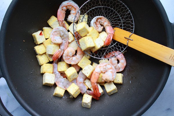 Shrimp and tofu cubes being fried in a wok with a slotted wire spoon inserted inside.