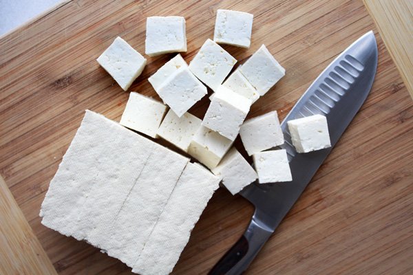 A block of tofu cut into cubes on top of a wooden cutting board with a chopping knife on the side.