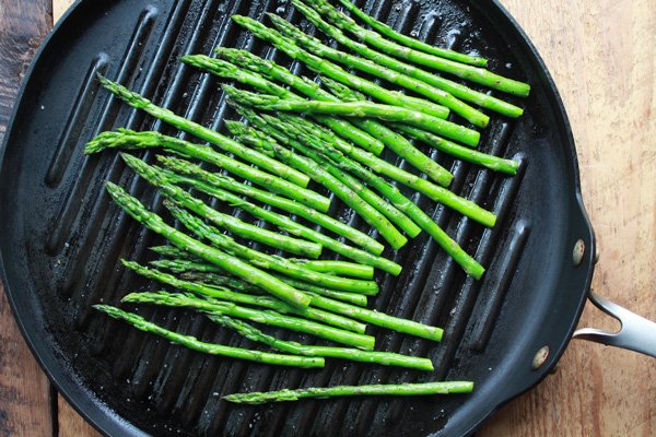 Asparagus spears being grilled on a round grill pan on top of a wooden board.