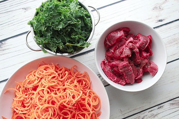 Three bowls filled with fresh green kale leaves, sliced sirloin beef, and spiralized sweet potatoes on top of a white plank board.