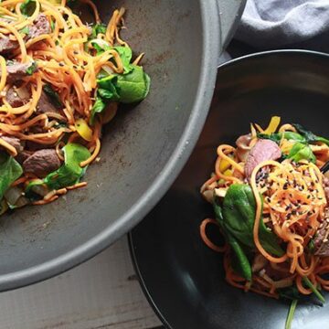 Stir-fried Korean sweet potato noodles with beef in a black bowl with a pair of black chopsticks and a wok filled with the stir-fried dish behind it.