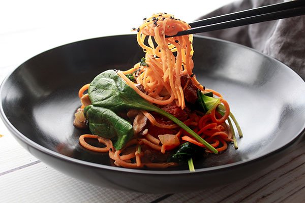 A black bowl filled with Korean sweet potato noodles with beef with a pair of black chopsticks lifting up spiralized sweet potato noodles above the bowl.