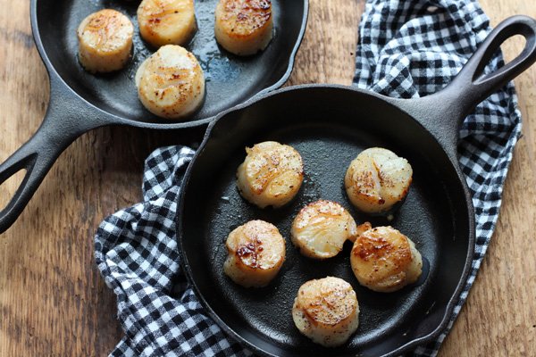 Two small cast iron pans with seared scallops inside on top of a checkered napkin on a wooden board.