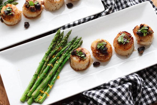seared scallops topped with a black bean sauce and a side of roasted asparagus on a long white plate on top of a checkered napkin