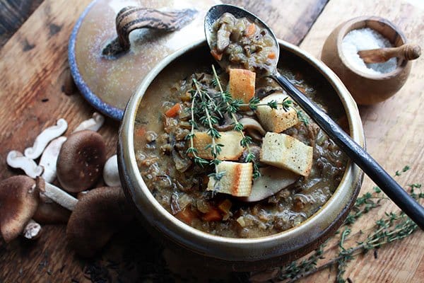A bowl of chunky wild rice mushroom soup topped with fresh croutons and sprigs of thyme on top of a wooden board with a large spoon on top and the soup lid on the side.