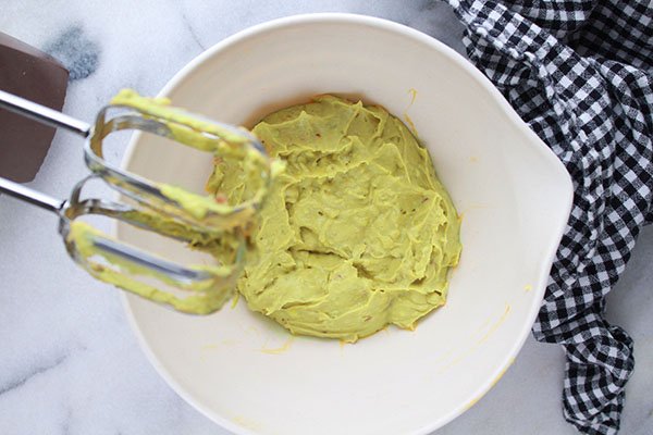 a hand blender beating avocado deviled eggs mixture in a white mixing bowl on top of a marble surface with a checkered napkin on the side