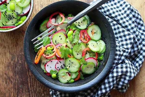 A black bowl filled with an Asian cucumber salad with lots of crunchy vegetables and sprinkled with black sesame seeds and topped with a fork with a black and white checkered napkin on the side.